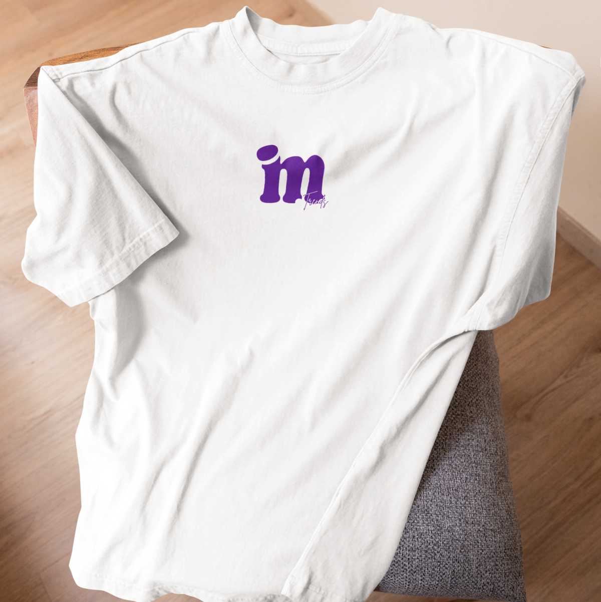 A white short-sleeve T-shirt laid flat on a chair over a wood floor, with a purple 'in' graphic printed on the chest.