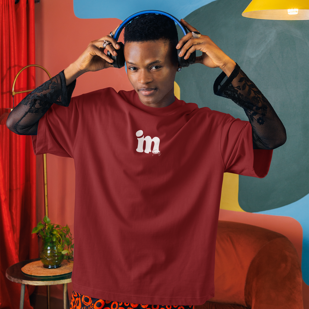 A person wearing a burgundy t-shirt with a white 'im' logo on the chest, layered over sheer black lace sleeves, and patterned orange and black shorts, while adjusting blue headphones in a colorful room with abstract wall art and a yellow lamp.