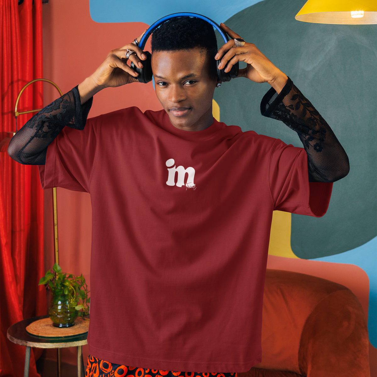 A person wearing a burgundy t-shirt with a white 'im' logo on the chest, layered over sheer black lace sleeves, and patterned orange and black shorts, while adjusting blue headphones in a colorful room with abstract wall art and a yellow lamp.